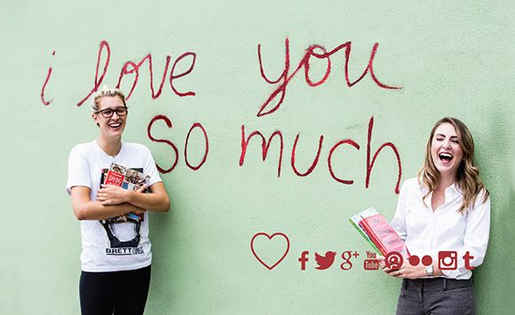 Two women holding books next to the "i love you so much' mural
