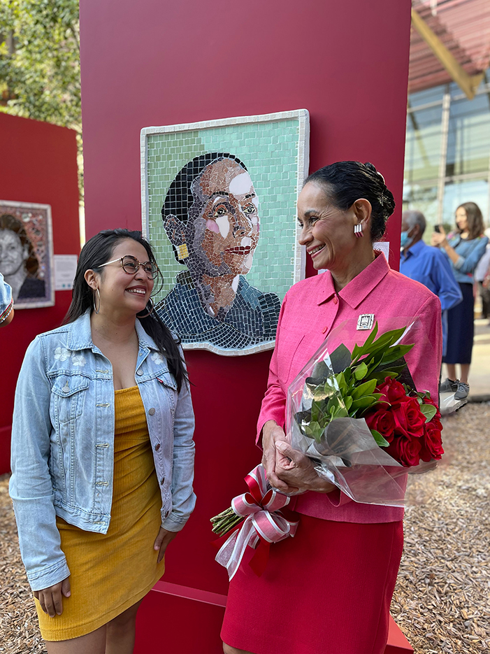 two women smiling in front of a portrait made with mosaic
