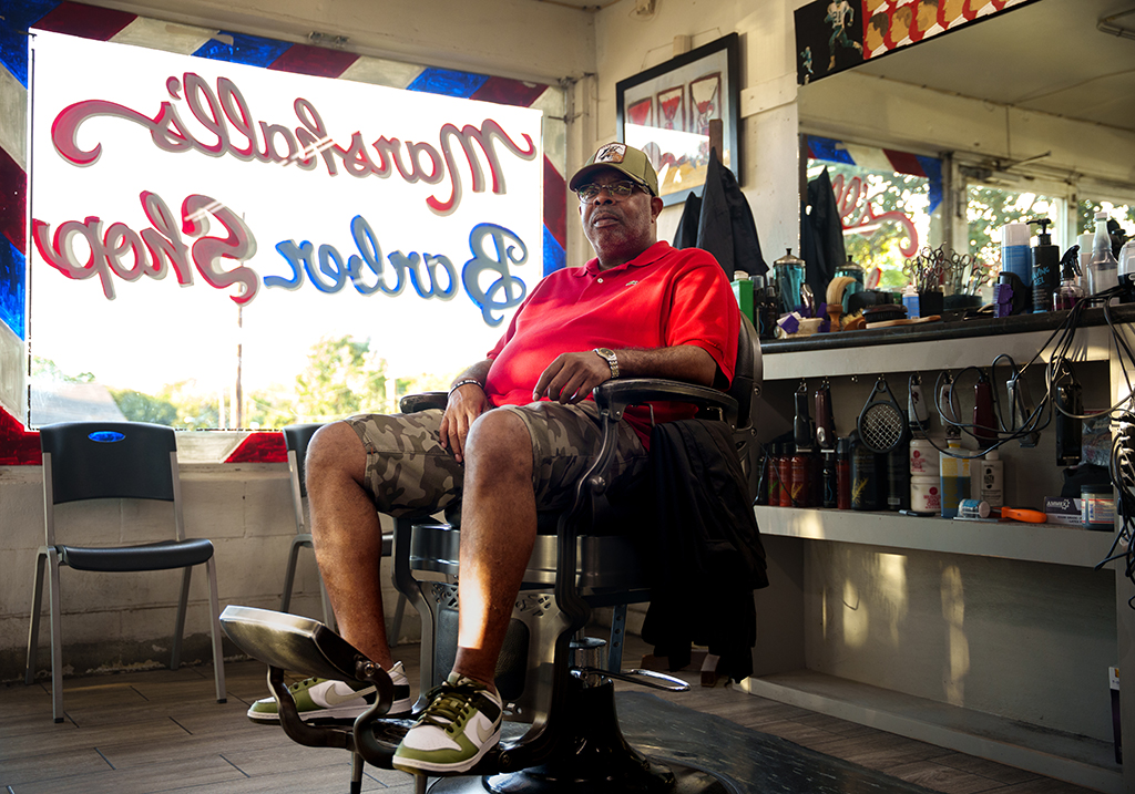 Black male seated in a barber chair, looking at camera, large text of barbershop in window