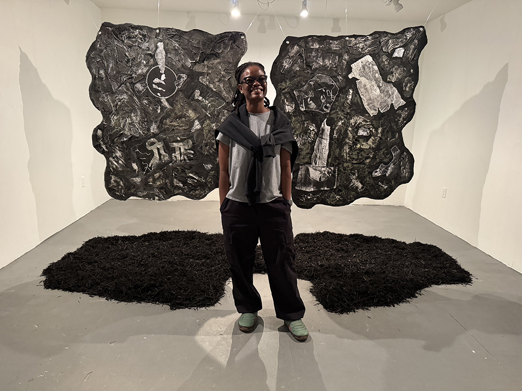 Black person standing in art gallery that has white walls, flag like art with grey abstract images hanging behind them, standing on abstract, carpet like shapes black carpet like s