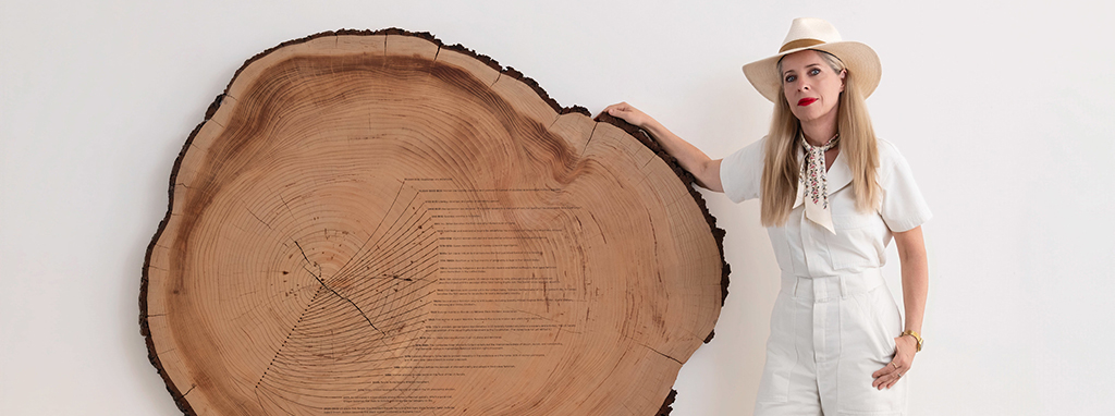 woman standing next to a cross section of a tree that's on a stand. Timeline of moments across women's history are etched into the tree ring
