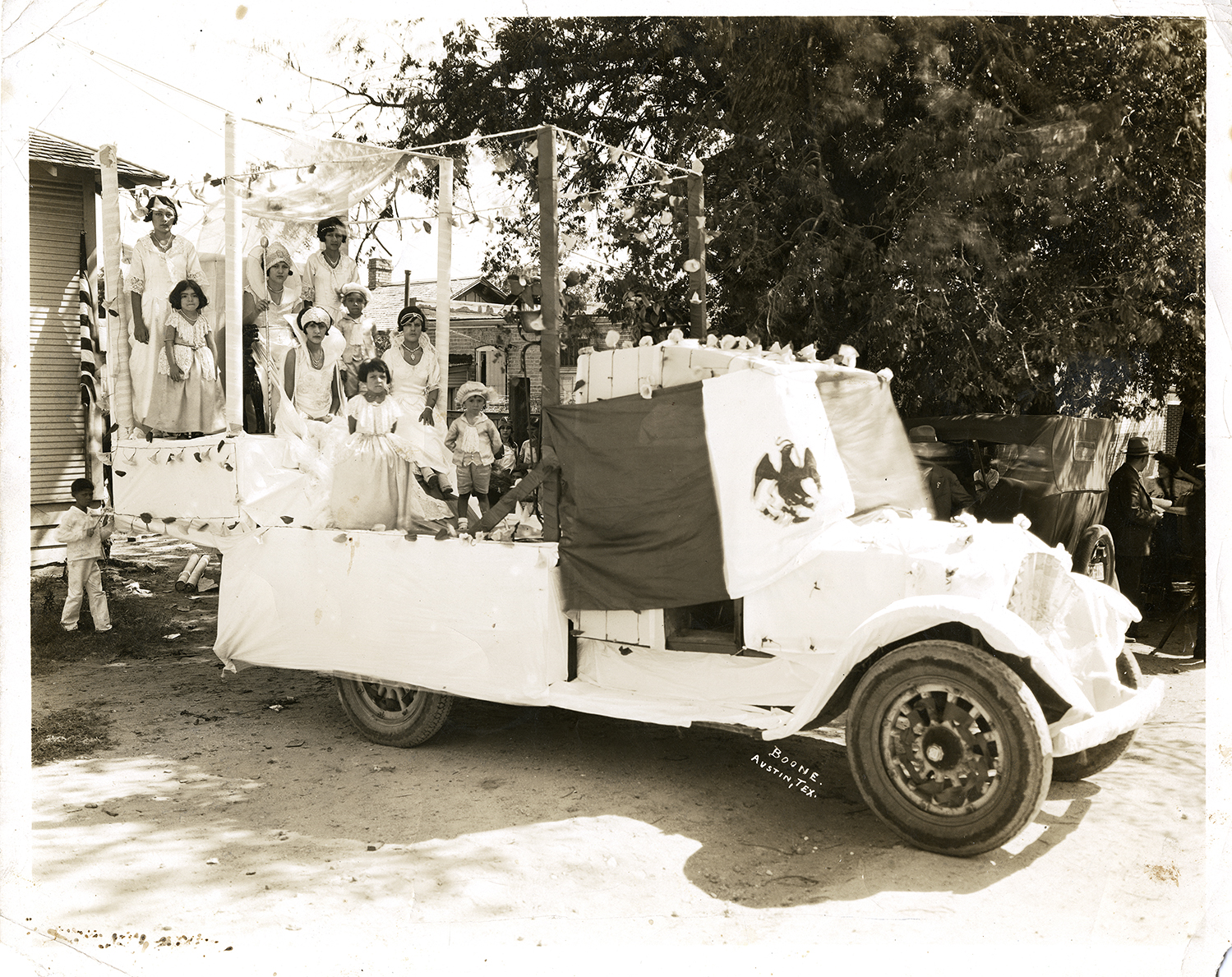 La Reyna de Dies y Seis along with her court stand in the bed of a truck which is draped with the Mexican flag and white sheets and adorned with paper flowers. Photo by Boone, PICA 36924