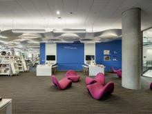 A white room with blue accent walls is broken up with curved tall white tables, white shelves of teen books, and pops of color with pink potato-chip shaped chairs