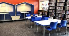 An orange wall with inset blue booth seating and two tables with blue chairs is capped off with a shelf of teen books. Signs on the tables read "Teen Space"