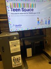 A PS5 gaming cabinet tells teens how they can ask to play videogames and sits next to a rolling TV cart says "Teen Space: this area is reserved for TEENS ages 13-18. Books are always available to all for browsing and borrowing."