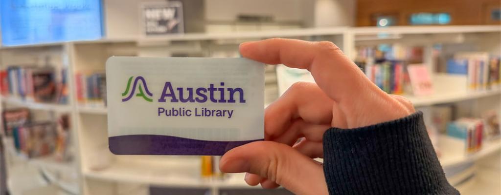 hand holding Austin Public Library card in front of book shelf