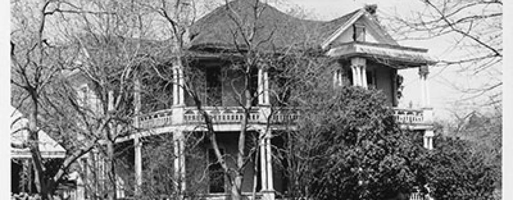 Close up, exterior view of two-story house on Avenue G. Surrounding landscape includes trees and shrubs. A roof-covered porch, with balcony and coulmns, surrounds each story. Neighboring house or building in partial view on the left.