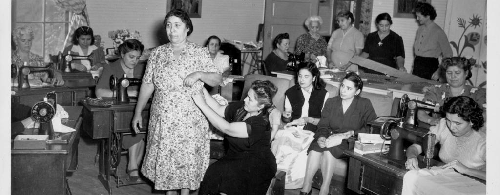 Fifteen women are in a room, some seated at sewing machines, some sitting and embroidering, others standing and measuring fabric. One woman wearing a floral patterned dress stands in the center of the room, while another woman in a black dress, sitting on a small bench, makes adjustments to the first woman's dress.