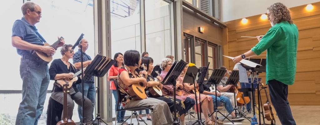 musicians seated, holding and playing their ukulele with a conductor at the front.
