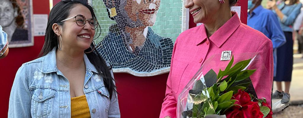 two women smiling in front of a portrait made with mosaic