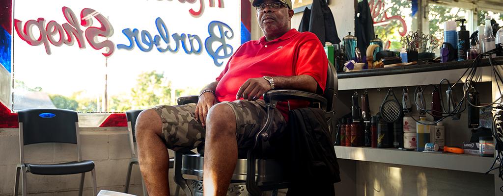 Black male seated in a barber chair, looking at camera, large text of barbershop in window