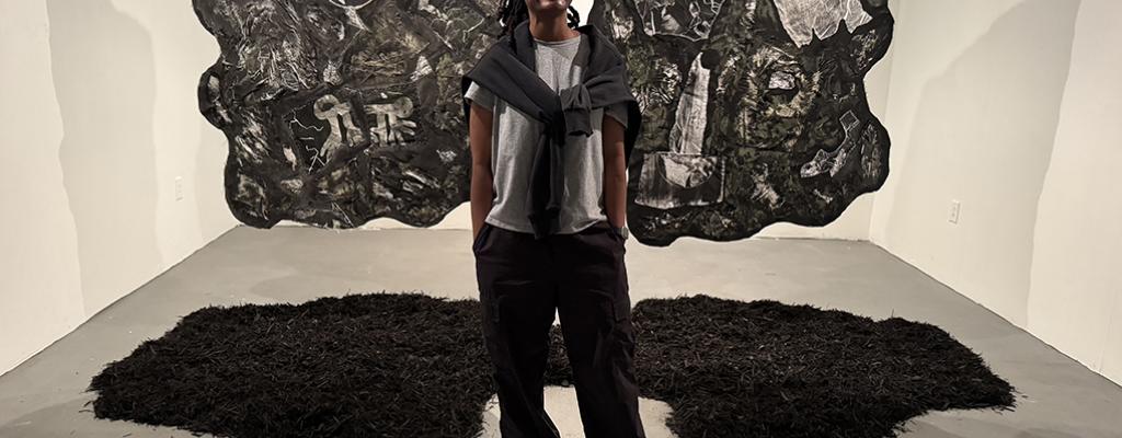 Black person standing in art gallery that has white walls, flag like art with grey abstract images hanging behind them, standing on abstract, carpet like shapes black carpet like s